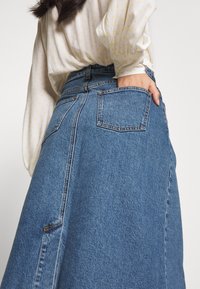 Džínová sukně v středně modré barvě s rovnou siluetou, kapsami, viditelným šitím a postranním rozparkem, spárována s lehkou blůzou s puntíky.