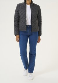 Grå quiltet jakke med opretstående krave og lynlås, kombineret med blå jeans og hvide sneakers, der viser et afslappet outfit.