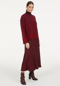 Frau trägt einen rot gemusterten Rollkragenpullover, einen dunkelroten Midirock und dunkelrote Ankle-Boots mit Blockabsätzen.
