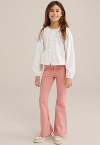 Vit långärmad blus med volangkant och knappstängning, parat med rosa utsvängda byxor och beige sneakers med texturerade detaljer.