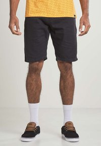 Black denim shorts with rolled cuffs, paired with white crew socks and black sneakers featuring brown accents. Model shown from waist down.