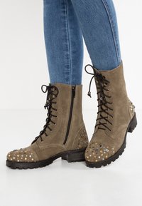 Olivově zelené semišové kotníkové boty s černým šněrováním, bočním zipem, hrotovou špičkou a robustní černou podrážkou.