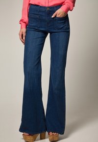 Mörkblå utsvängda jeans med hög midja, som har framfickor och knappstängning. Texturen är slät denim. Bärs med korkplattformshälar.