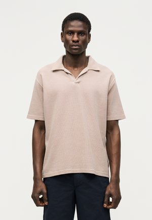 Man draagt een lichtbeige korte mouw polo en donkere broek, staand voor een effen lichtgrijze achtergrond.