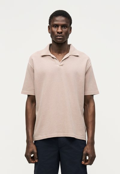 Homme portant un polo beige clair à manches courtes et un pantalon foncé, debout devant un fond gris clair uni.