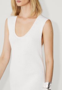 Robe tricotée blanche sans manches avec un profond décolleté en V, présentant un motif texturé et une silhouette ajustée, complétée par un bracelet métallique.