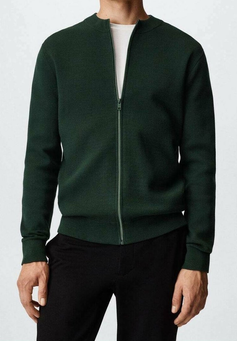 Homme portant un cardigan zippé vert foncé sur un t-shirt blanc et un pantalon noir, montré du cou jusqu'aux hanches.