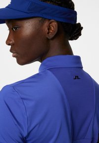 Blå atletisk polo med nätpanel på baksidan och svart logodetalj, tillverkad av lätt, fukttransporterande material.