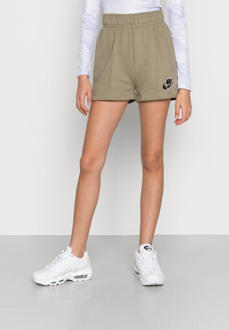 Shorts en coton vert olive avec ceinture élastique, poches latérales et logo Nike noir sur la cuisse gauche. Portés avec des baskets blanches.
