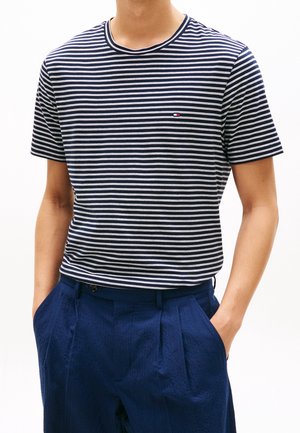Man draagt een marineblauw-wit gestreept kortemouwenhemd, ingestopt in geplooide marineblauwe broek, met handen in de zakken.