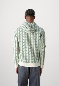 Felpa con cappuccio in crema con un motivo geometrico verde. Presenta una tasca frontale a marsupio e polsini a coste. Tessuto morbido, vestibilità rilassata.