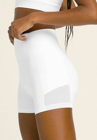Donna che indossa dei pantaloni a compressione bianchi a vita alta con pannelli in rete, mostrata di lato, mentre sistema il cinturino con la mano.