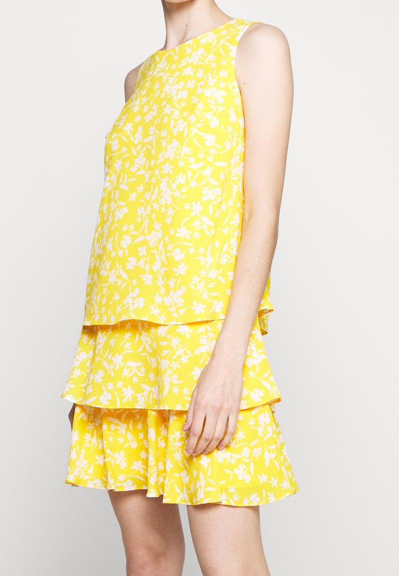 Gul blommig klänning med ärmlös design, som har lager av volanger. Tyget har en strukturerad yta med vita blommönster.
