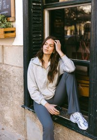 Grijze zip-up hoodie, geribde grijze broek en witte sneakers. Model zit bij een raam, houdt een koffiekop vast, met een achtergrond van een gestructureerde stenen muur.