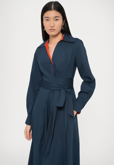 Granatowa sukienka kopertowa z długimi rękawami, dużym kołnierzem i pomarańczowymi akcentami. Wykonana z gładkiej tkaniny, z przewiązanym w talii pasem i guzikami z przodu.