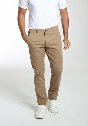 Homme portant un chino beige, un t-shirt blanc et des baskets blanches, debout avec une main dans la poche sur un fond uni.
