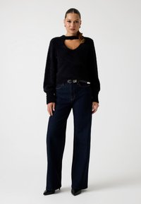 Jersey negro texturizado con un diseño de ojal, combinado con unos jeans de mezclilla oscuros de tiro alto y zapatos negros de punta. Estilo simple y moderno.