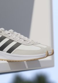 Białe i szare sportowe buty z czarnymi paskami i gumowymi podeszwami, wystawione na białej platformie na tle szaro-niebieskim.