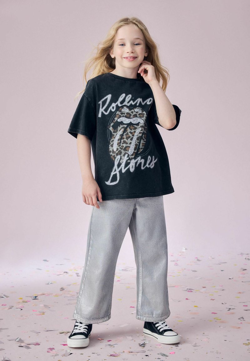 Child with long blonde hair wears oversized black Rolling Stones t-shirt, wide silver pants, and black sneakers standing on confetti-covered floor.