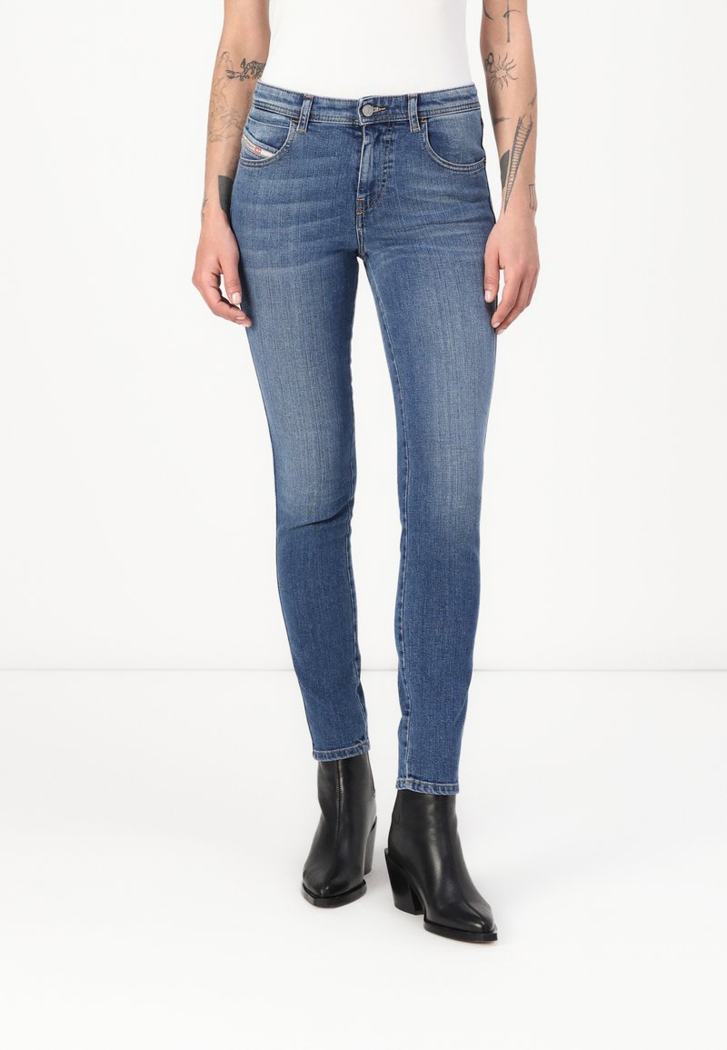 Vaqueros de denim azul ajustados con un ligero desgaste, cinco bolsillos y una cintura de altura media. Llevados con botas de tobillo negras. Textura lisa.