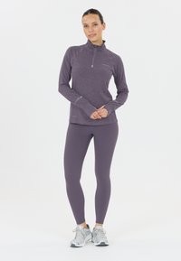 Kvinna står med händerna i kors, klädd i en lila långärmad zip-up-tröja, matchande leggings och silverfärgade löparskor på en vit bakgrund.