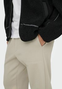 Black fleece jacket with textured fabric, silver zipper, and contrasting seams. Beige trousers with smooth texture and side pockets.