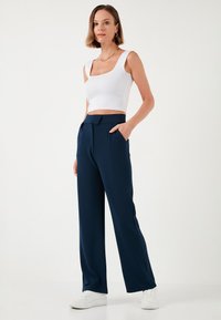 Crop top blanc à larges bretelles associé à un pantalon large bleu marine avec une ceinture et des poches latérales, porté avec des baskets blanches.