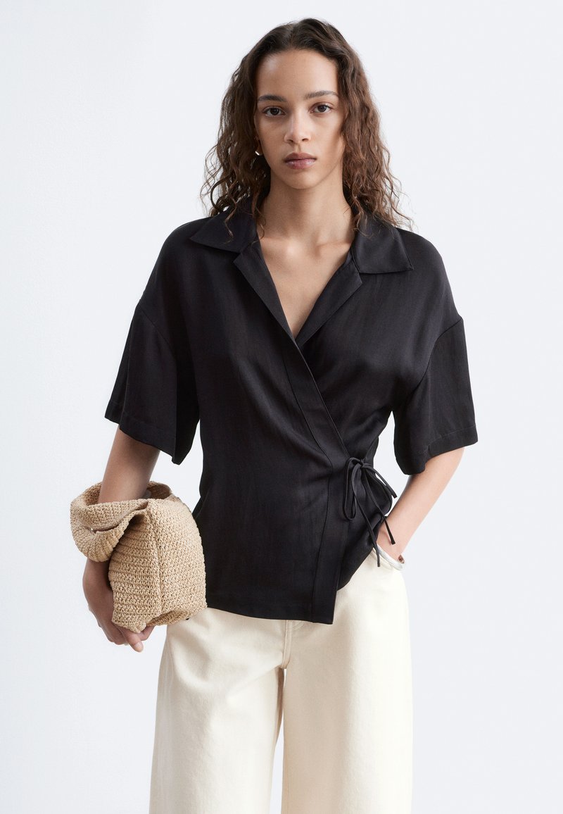 Femme aux cheveux bouclés porte un top cache-cœur noir et un pantalon blanc, tenant un sac tressé beige sur un fond clair uni.