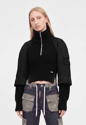 Femme aux cheveux blonds raides porte un pull court noir avec un col zippé haut et un jean foncé avec de grandes poches cargo grises.
