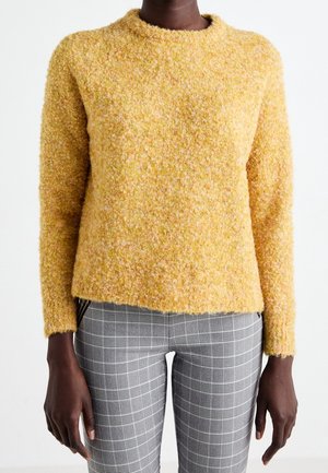 Osoba nosi żółty sweter o fakturze z długim rękawem oraz szare spodnie w kratę, stojąc na tle jednolitego białego tła.