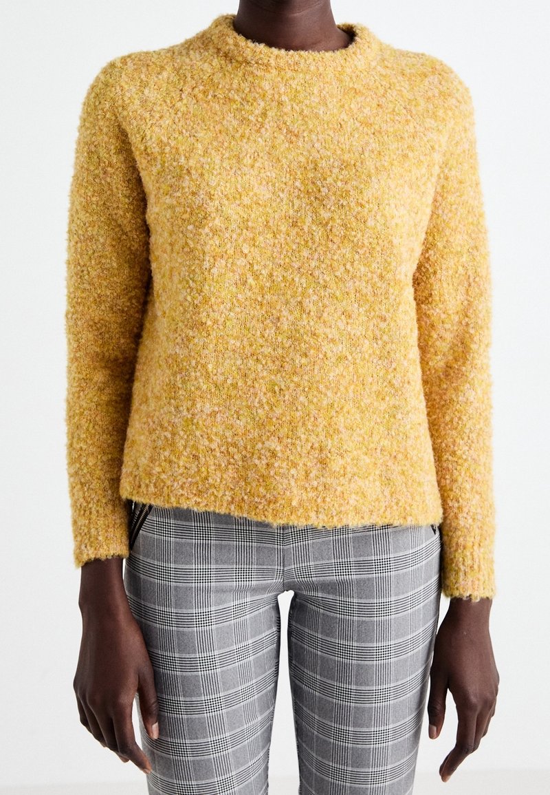 Une personne porte un pull jaune texturé à manches longues et un pantalon à carreaux gris, debout devant un fond blanc uni.