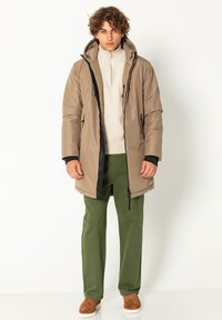 Beige isolierte Jacke mit hohem Kragen, schwarzen Reißverschlüssen und Seitentaschen, kombiniert mit grünen Hosen und braunen Slippern.