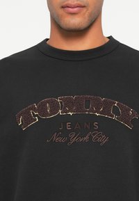 Čierna mikina s vyvýšeným, textúrovaným logom "TOMMY" v tmavohnedej farbe a zlatým obrysom, s nápisom "JEANS" a "New York City" vytlačeným nižšie.