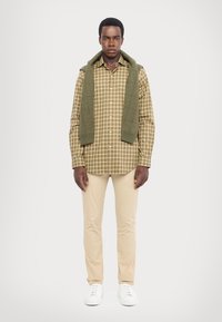 Checkered brown and green shirt, beige pants, and white sneakers; green sweater draped over shoulders. Smooth fabric with a relaxed fit.