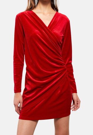 Kobieta ubrana w czerwoną aksamitną mini sukienkę z długim rękawem, z kopertowym fasonem i marszczeniem po bokach, uzupełnioną długimi srebrnymi kolczykami.