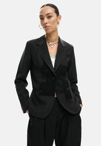 Blazer nero a righe sottili con pannelli neri a contrasto, chiusura frontale con un solo bottone e rever a lancia. Abbinato a pantaloni larghi abbinati.