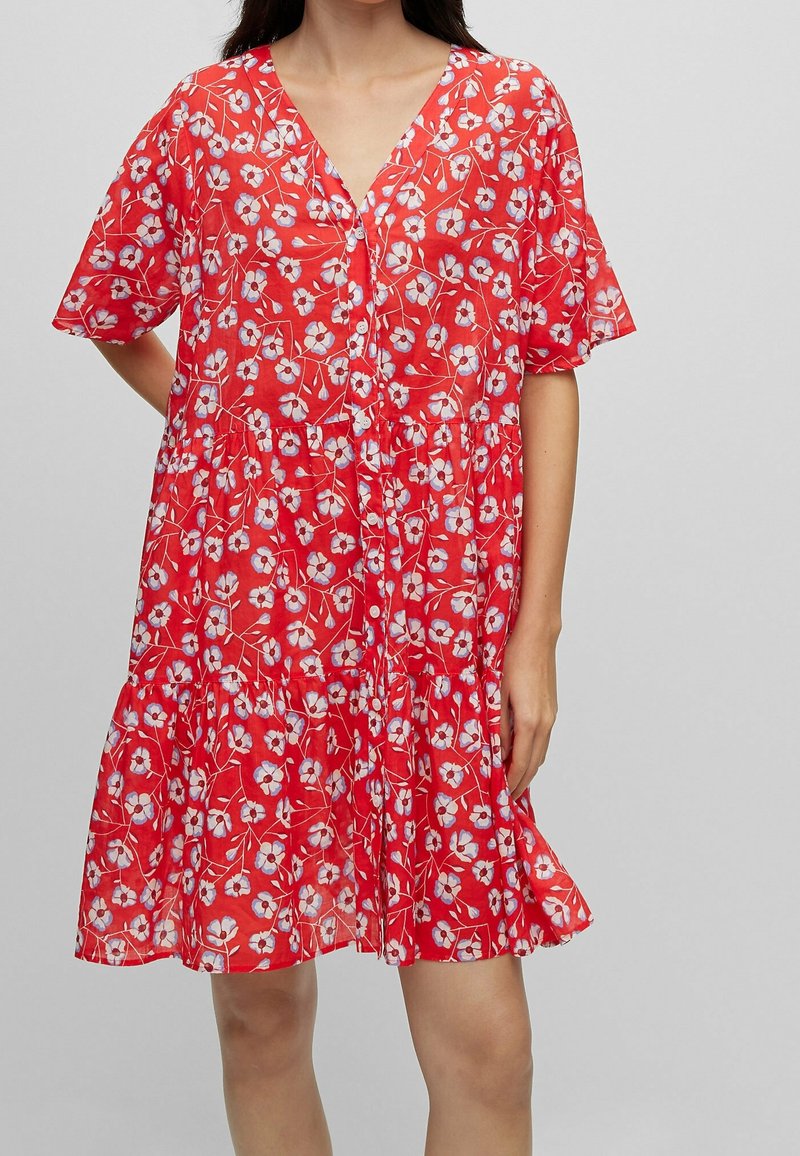 Femme portant une robe rouge ample à manches courtes avec un motif floral blanc et violet, debout devant un fond uni.