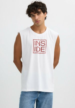 Joven con cabello rizado y oscuro, vistiendo una camiseta blanca sin mangas con texto rojo "INSIDE" y jeans azules, frente a un fondo liso y claro.