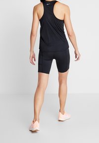 Zadní pohled na osobu v černém bezrukávovém atletickém topu, černých cyklistických šortkách s bočními kapsami a světlých růžových běžeckých botách.