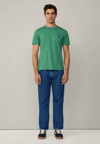 Zelené bavlnené tričko s okrúhlym výstrihom a krátkymi rukávmi, ozdobené modrým lemom na golieri a potlačeným číslom „2“ na ľavej strane hrudníka. Dopĺňané modrými džínsami.