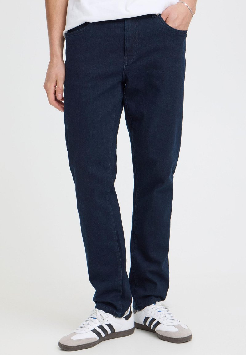 Personne portant un jean bleu foncé, des baskets blanches avec des bandes noires, une main dans la poche, debout devant un fond uni.