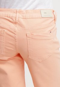 Pantalones de denim color durazno con un diseño de tiro medio, que cuentan con dos bolsillos traseros y detalles de costura sutiles. La tela tiene una textura suave.
