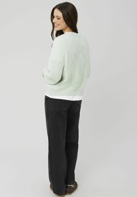 Pull en grosse maille vert clair avec un col rond, superposé à une chemise blanche, associé à un pantalon large noir et des chaussures marron.