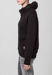 Felpa con cappuccio in cotone nero con cappuccio piegato a strati e zip laterale. Presenta polsini e orlo a costine e una piccola toppa con logo colorato sul davanti.