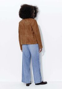 Veste en daim marron avec une coupe simple, associée à un pantalon en denim bleu clair à jambes larges doté de boutons sur les côtés et de chaussures noires.