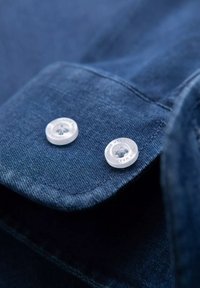 Polsino di camicia in denim con due bottoni in plastica trasparente. Il tessuto presenta una texture liscia e un colore blu profondo, evidenziando i dettagli della cucitura.