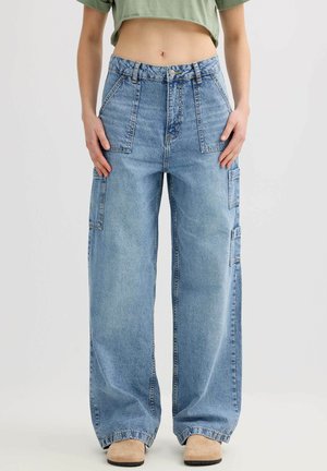 Persona que lleva pantalones cargo vaqueros de pierna ancha y cintura alta en azul claro, con grandes bolsillos delanteros y laterales, combinados con zapatos beige y una camiseta corta verde.