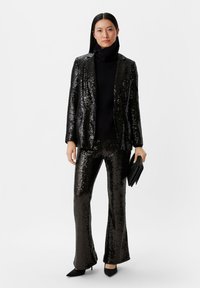 Zwarte paillettenblazer en bijpassende flared broek, met een hooggesloten gebreid top, zwarte clutch en schoenen met spitse neuzen. Gestructureerde, glanzende afwerking.