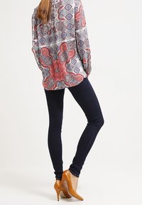 Langärmlige gemusterte Bluse in Rot, Blau und Weiß mit Paisleymuster, kombiniert mit dunkelblauen Skinny-Jeans und hellbraunen High Heels.
