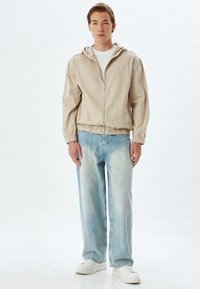 Beige vuorilla varustettu vetoketjullinen takki, jossa on joustavat hihansuut ja helma. Vaaleansiniset väljyysfarkut, joissa valkoiset lenkkarit. Minimalistinen muotoilu.